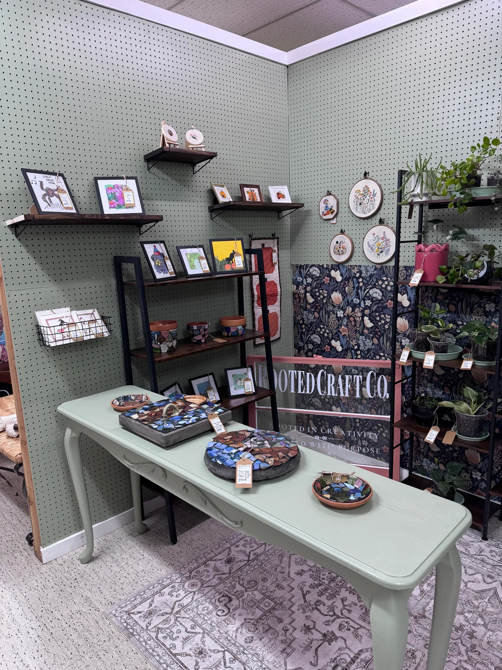 Rooted Craft Co booth with plants, mosaics, framed art, embroidery, and quilts